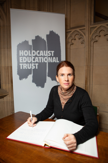Trudy wearing a black cardigan, brown polo neck knitted top, signing the book of commitment in front of a Holocaust Education Trust banner
